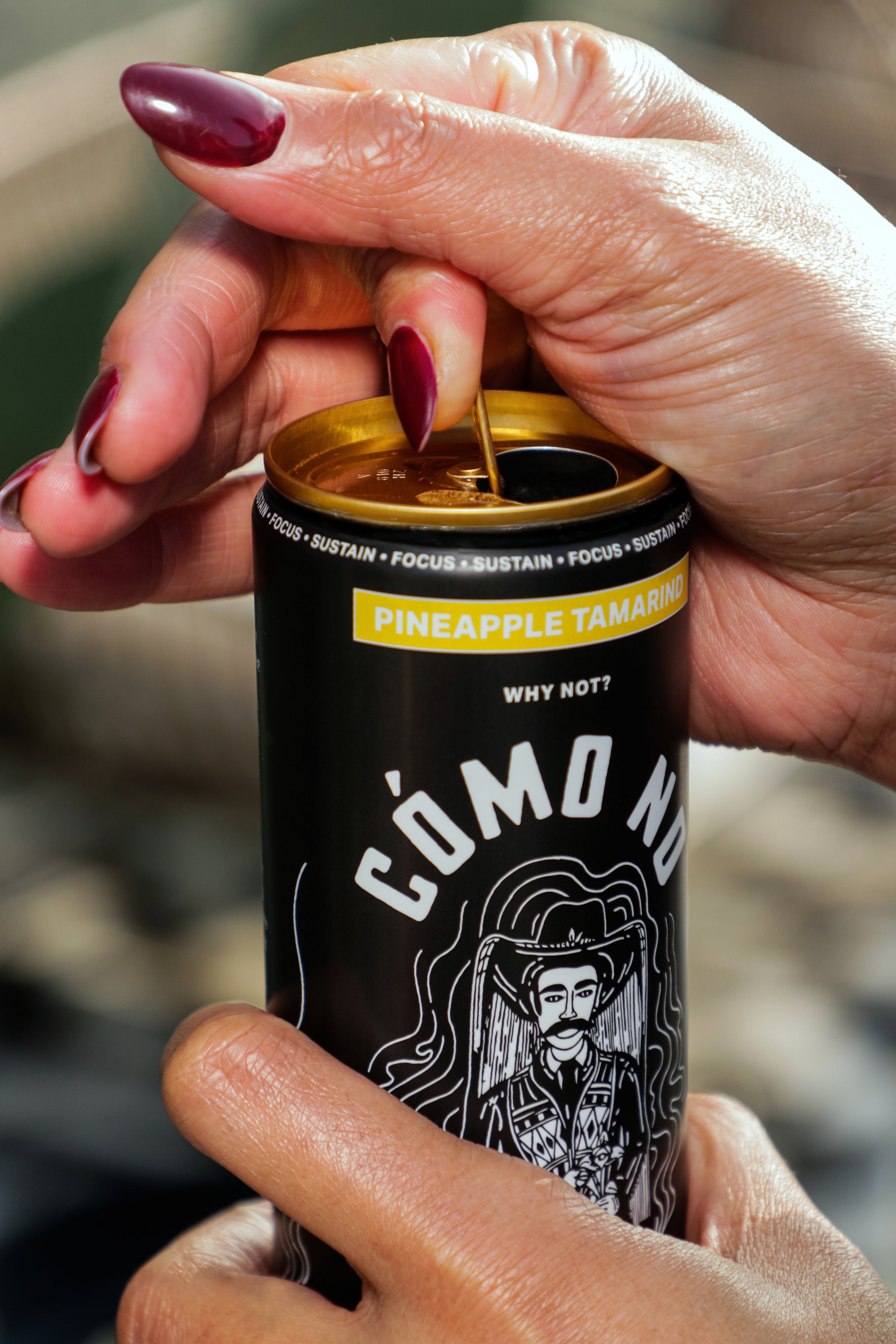 Hand opening a can of 12 Pack Pineapple Tamarind Como No energy drink, showing black can with gold accents and white logo